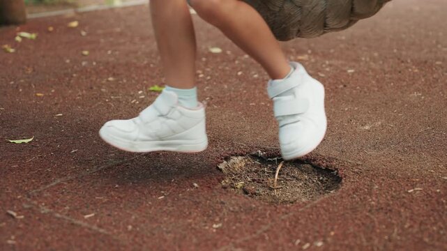 Girl stepping near playground surface hole, white sneakers scuffing rubber crumb ground, braided rope swing overhead, curious toes testing texture, potential safety concern, candid park outing moment