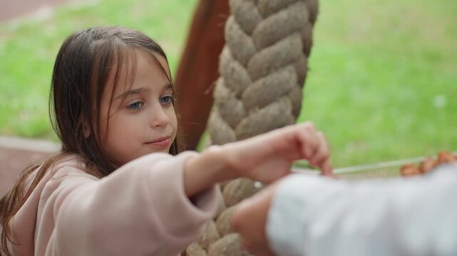 White daughter takes cookie from mom on rope swing in park reaching hand meets warm chocolate cookie, candid smile, crumbs on fingers, cozy hoodie, gentle outdoor light, wholesome family bonding