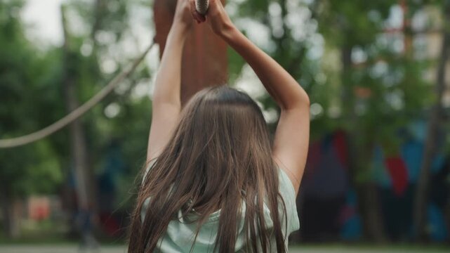 Daughter climbing rope course from behind, long hair falling over shoulders, hands gripping high rope and pulling with steady resolve, timber posts and green canopy backdrop, adventurous training vibe