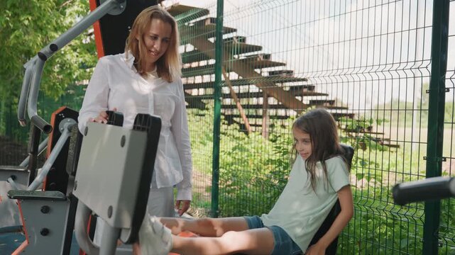 White mom and daughter at fitness machine, mother guides daughter to place feet and try seat, patient instruction in leafy park, attentive expressions, safety minded family learning moment