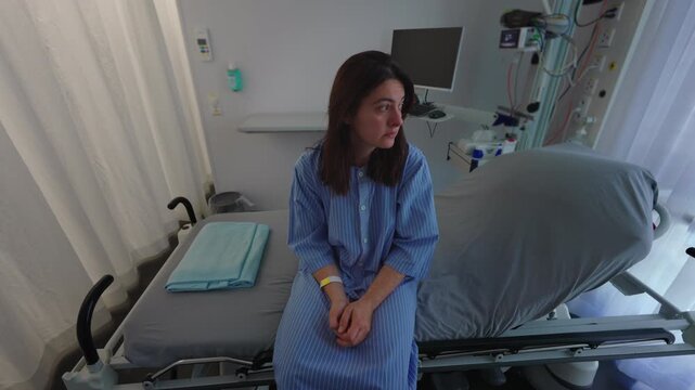Female patient sitting on hospital bed in medical room looking aside with worried expression reflecting anxiety uncertainty and emotional fatigue during recovery