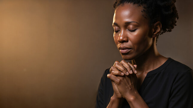 Black woman praying with eyes closed and clasped hands. Religious faith and spirituality concept. Studio portrait with copy space on dark background