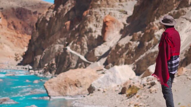 Rack focus shot of an Indian male tourist standing in front of blue colored Tsarap river flowing in the middle of Himalayas as seen in Zanskar Valley of remote Ladakh, India. Scenic background.