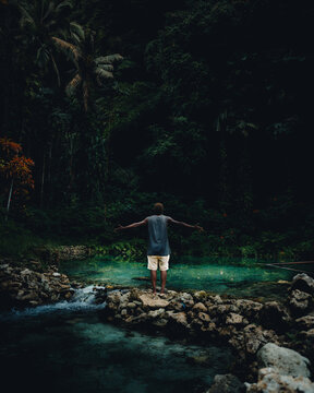 View of a lone figure stands poised on the rocky edge of a turquoise pool, arms outstretched, embraced by the emerald depths of the surrounding forest, Lata, Solomon Islands.