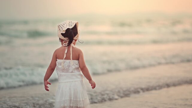 Adorable toddler girl in white dress standing on a peaceful beach at sunset, looking at the ocean waves. A cinematic back view capturing childhood wonder, wellness, and freedom during a family summer