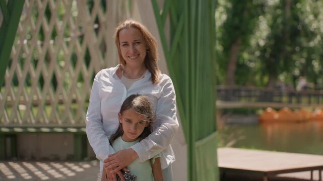 Mom and daughter portrait at lakeside gazebo, mother gently hugging child, warm sunlight filtering through trees, calm water with orange paddleboats in background, soft summer greenery, candid tender