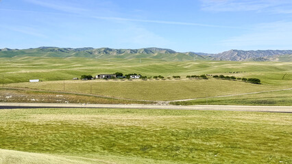 Obraz premium Breathtaking view of a small rural town nestled among lush green meadows and rolling hills in California. A scenic road leads to the village under a clear blue sky and majestic mountains.