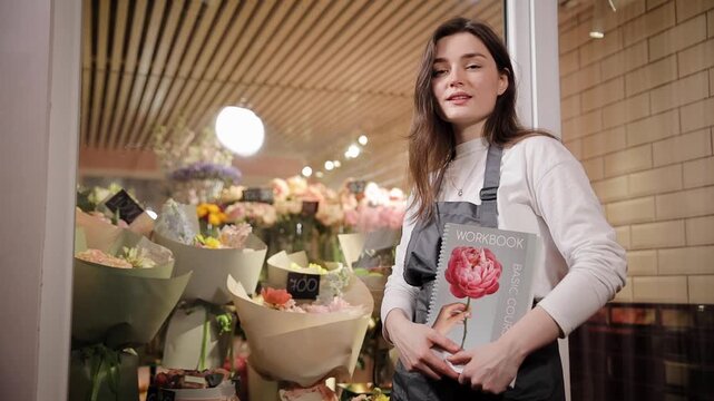A young woman in a flower shop holding a basic course workbook, wearing an apron, teaching skills related to floristry amidst a lively and inspiring environment