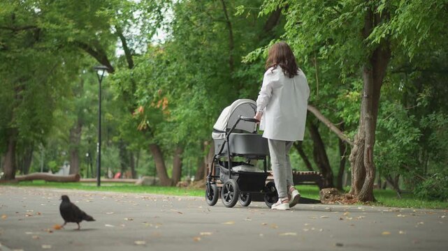 Caucasian mother stops by crow in park, observant pause beside stroller near bench, leafy canopy overhead, cautious protective stance, urban green path, soft overcast light, attentive parental mood