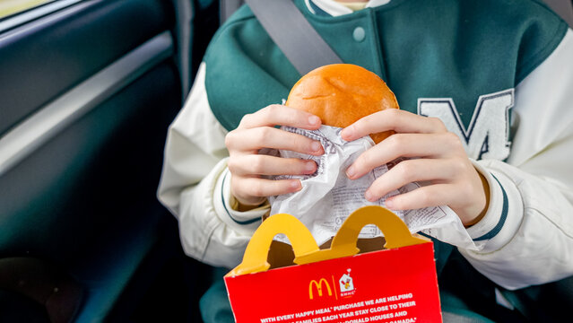 Child holding McDonald's hamburger in car, Happy Meal box visible, wearing green and white varsity jacket with letter