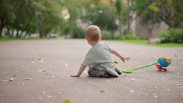 Toddler sitting on pavement with toy truck, reaches and explores colorful wheels, candid outdoor play scene showing curiosity, early motor development and parental lifestyle routine.