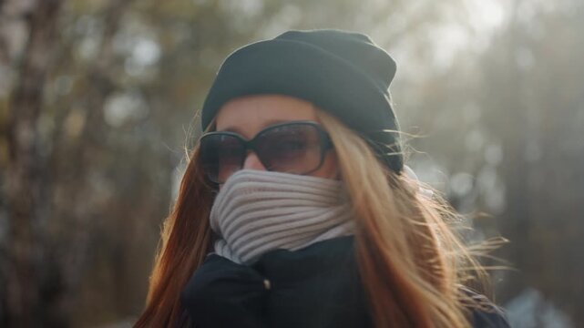 Masked white woman in forest backlit by autumn sun, beanie and scarf hiding lower face, sunglasses and backpack, slow cautious walk suggesting prowler or snatcher, cinematic moody tension with closeup