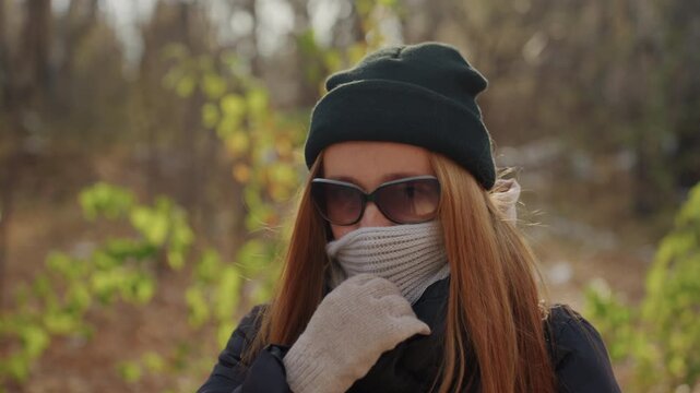 White woman smiling under knit hat adjusting scarf with gloved hand, sunglasses, backpack straps visible, autumn sunlight through leaves, playful yet furtive expression, candid outdoor moment