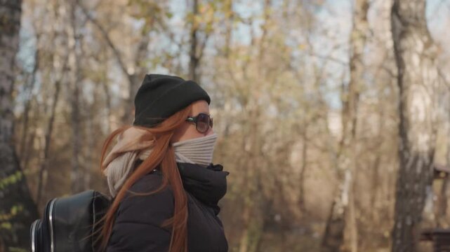 Caucasian woman side profile on trail wearing beanie and scarf, carrying black backpack resembling instrument case, auburn hair blowing in breeze, sunglasses and steady pace through birch woods