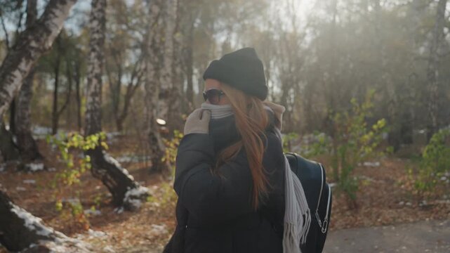 White woman backpacker walking forest trail, masked and glancing over shoulder under autumn sunlight birch grove setting, cautious pace and tense posture hinting at prowler presence and snatcher