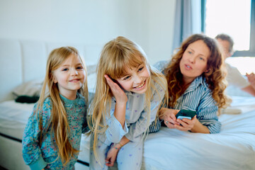 Obraz premium Happy family with parents and daughters spending morning time in bedroom, mother showing something on smartphone to children