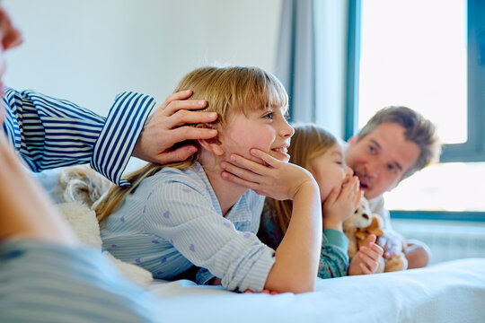 Happy family spending time together in bed, a child listening while a parent gently touches head, another child whispering