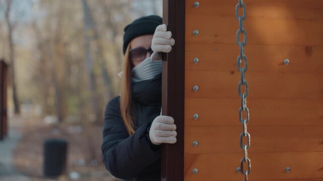 Masked caucasian woman peeking behind panel, outdoor park scene with wooden panel and chain, gloved hands grip edge, sunglasses and scarf conceal face, furtive motion suggests prowler snatcher intent,