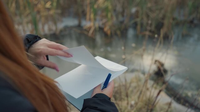 Lady checking lengthy documents closely. Female inspecting and flipping through sheets carefully. Closeup shot of woman closely analyzing and turning through multiple paper sheets beside marsh area