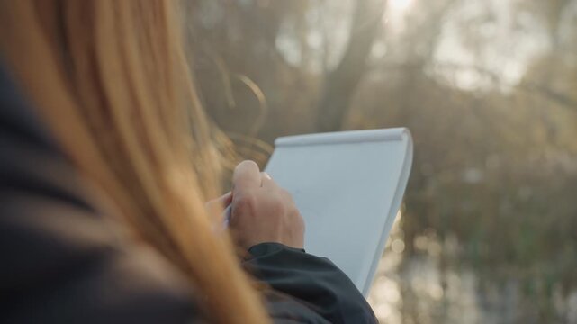 Lady captures notes beside flowing water. Caucasian woman jotting quick sketches in peaceful outdoor setting. Woman with long hair sketches and notes beside river during tranquil golden sunset