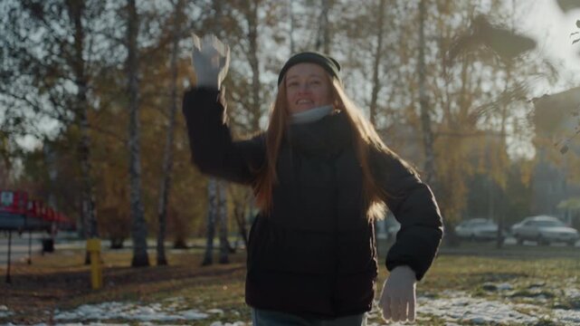 Sunlit winter park action white woman, caucasian woman throws snowball with laughter, mittened hands release powdery snow, dynamic midair motion, birch trees and scattered snow on grass, upbeat