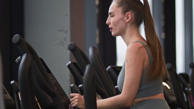 Medium shot of young Caucasian woman with long ponytail doing cardio on elliptical trainer in gym