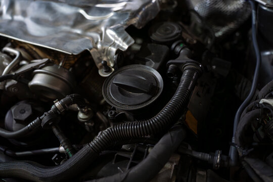 Close view of a car engine bay with oil filler cap, hoses, wiring, and metal components under the hood in workshop lighting.