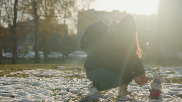 Backlit woman crouching over toy in snowy urban park at dusk, hooded backpack, furtive reach like snatcher retrieving small doll, warm lensflare and long shadows, cold atmosphere, cinematic tension