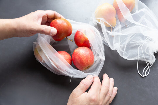 Close-up of hands holding a red apple and a white textile grocery bag on a dark background. Concept of zero waste lifestyle, sustainable shopping, and plastic-free environment.
