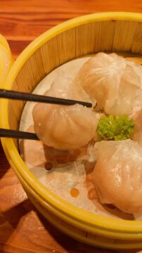 Hand picking a translucent har gow shrimp dumpling with chopsticks from a steaming bamboo basket, showcasing classic cantonese dim sum ready to enjoy at the table
