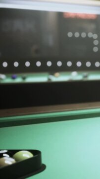A group of pool balls sits on a green table. People play pool in a game area at night. The atmosphere has bright lights and the sound of balls hitting each other.
