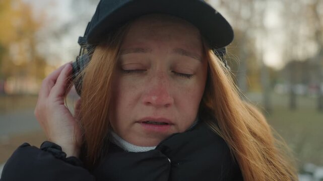 Closeup redhaired woman with headphones, tearful eyes and runny nose, shivering in puffy coat while gripping stuffed elephant, listening for prowler or snatcher in blurred autumn park, intimate