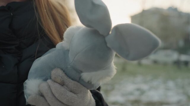Young white woman clutching gray bunny, mittened hands protecting plush in winter park, tense expression and guarded posture, soft fur and puffy jacket visible, faint snow on grass, urban buildings