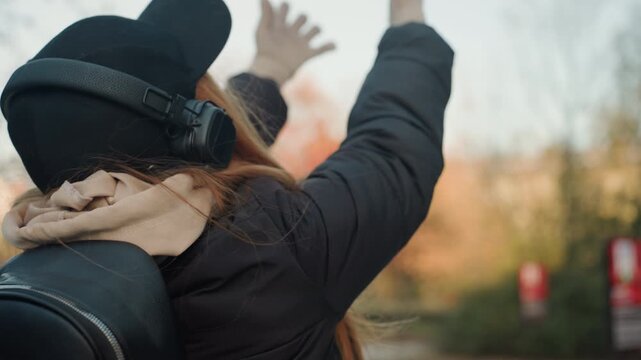 Woman backpack headphones autumn park reacting to suspected snatcher attempt, reaching toward guitar case, wearing cap scarf coat, candid back and side profile, trees and fall leaves in soft bokeh,