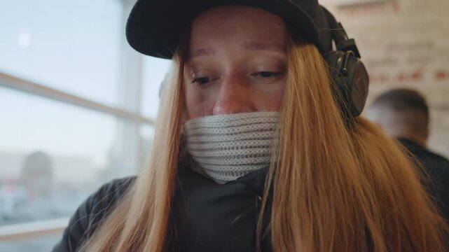 White redhead woman covering face with knit scarf and headphones by window, pulling scarf up with gloved hands inside cafe tense eyes scan surroundings, furtive body language, cap and winter coat