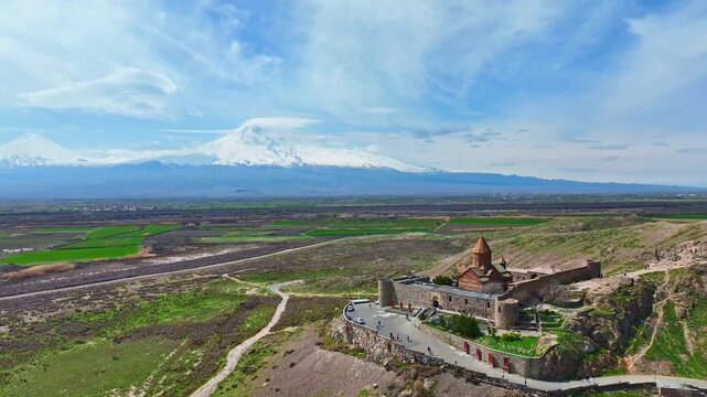 Aerial view shows ancient Khor Virap monastery perched on rocky hill. Snow-capped mountains rise behind vibrant green fields. Clear blue sky with wispy clouds adds calm beauty