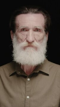 Vertical view of senior Caucasian man with long beard facing camera. Male keeping neutral expression and steady gaze. Showing age lines and wisdom. Standing in dark studio background.