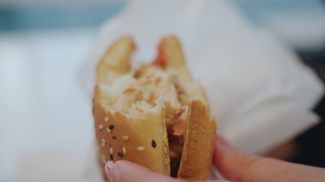 Closeup hand grabbing burger in cafe, patron snatches quick bite from wrapped sesame bun, creamy sauce and chicken patty visible, shallow focus on fingers and food, soft natural light, urban snack