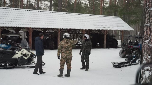 Crew gathers by atv shelter, winter basecamp with snowmobiles and gear, camo rider converses with guide, men in helmets and jackets, pine forest background, exchanging route plan, friendly briefing,