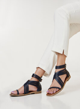 Young woman modeling stylish Mediterranean-inspired leather gladiator sandals in dark blue, paired with white cropped trousers. The scene evokes quiet luxury, emphasizing minimalist, sophisticated.