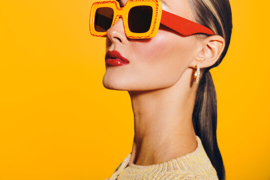 Stylish woman with slicked back hair wears large orange sunglasses and red lipstick against bright yellow background. Concept of fashion, beauty, and summer style.