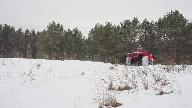 Red atv approaching across snowy field. White rider wearing camouflage and helmet drives steadily toward camera, tire tracks cutting powder, pine tree backdrop, closeup frontal view highlighting