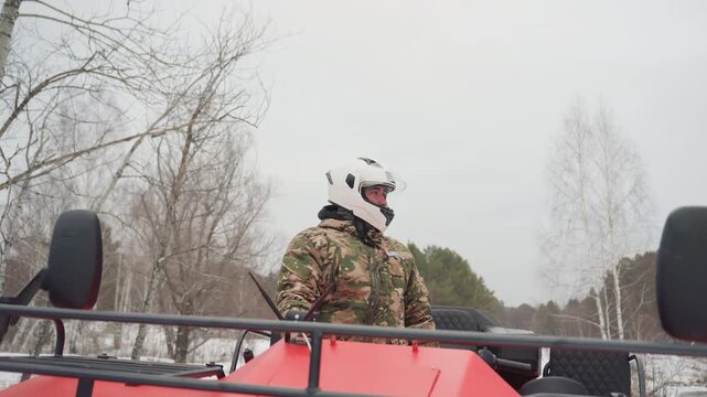 Caucasian rider scouting snowy trail on red atv, wearing camouflage jacket and white helmet, surveying terrain and horizon, readying for extreme trail run, alert posture and handheld radio visible,