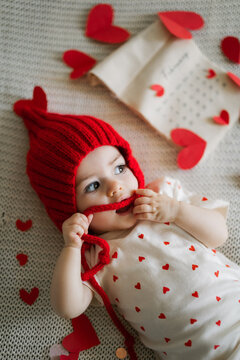 Little baby girl in red knit hat smiles while holding string near heart shaped decorations on soft textured surface