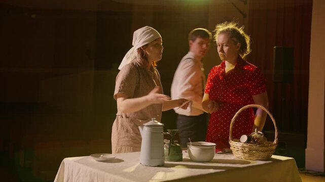 Two women performing an emotional kitchen scene on stage, exchanging dialogue over a table with dishes and basket props under warm theatrical lighting