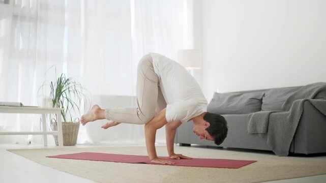 Athletic man performing crow yoga pose on a mat in a bright modern apartment. Young male, person or adult balancing on his hands during an advanced home workout, yoga practice or wellness routine in a