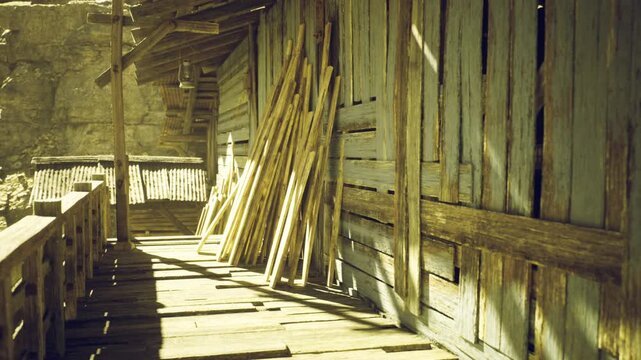 Sunlight filters through a rustic village as wooden boards lean against the weathered wall. The scene evokes a sense of history and craftsmanship in this quiet, timeless space.