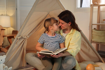 Mother and her daughter reading book near toy wigwam at home © New Africa