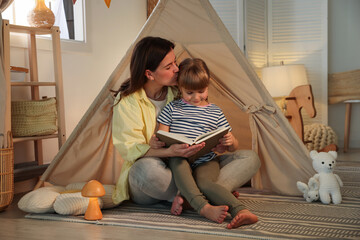 Mother and her daughter reading book near toy wigwam at home © New Africa