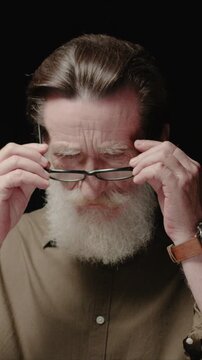 Vertical view of adult Caucasian man putting on glasses and looking down. Male adjusting eyewear with both hands. Focusing on vision and reading. Standing in dark background in studio.